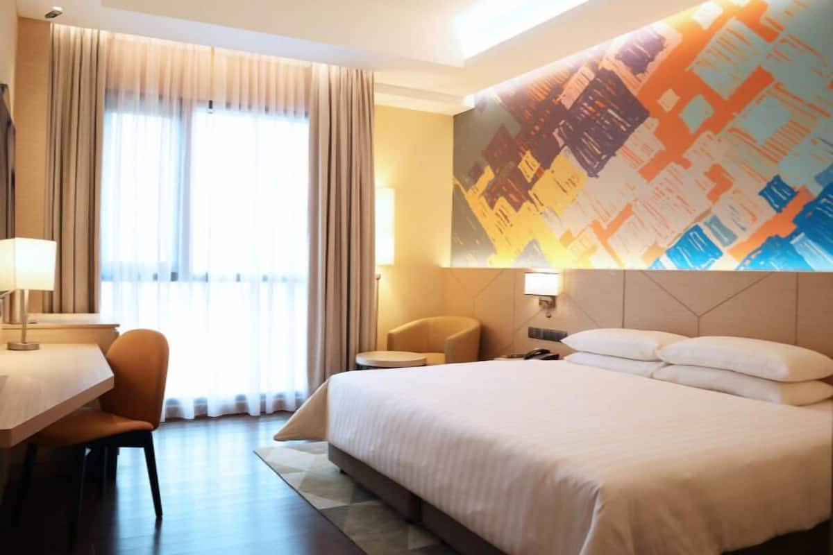 Habitación de hotel con cama, escritorio, silla, ventana grande con cortinas y un mural colorido en la pared.