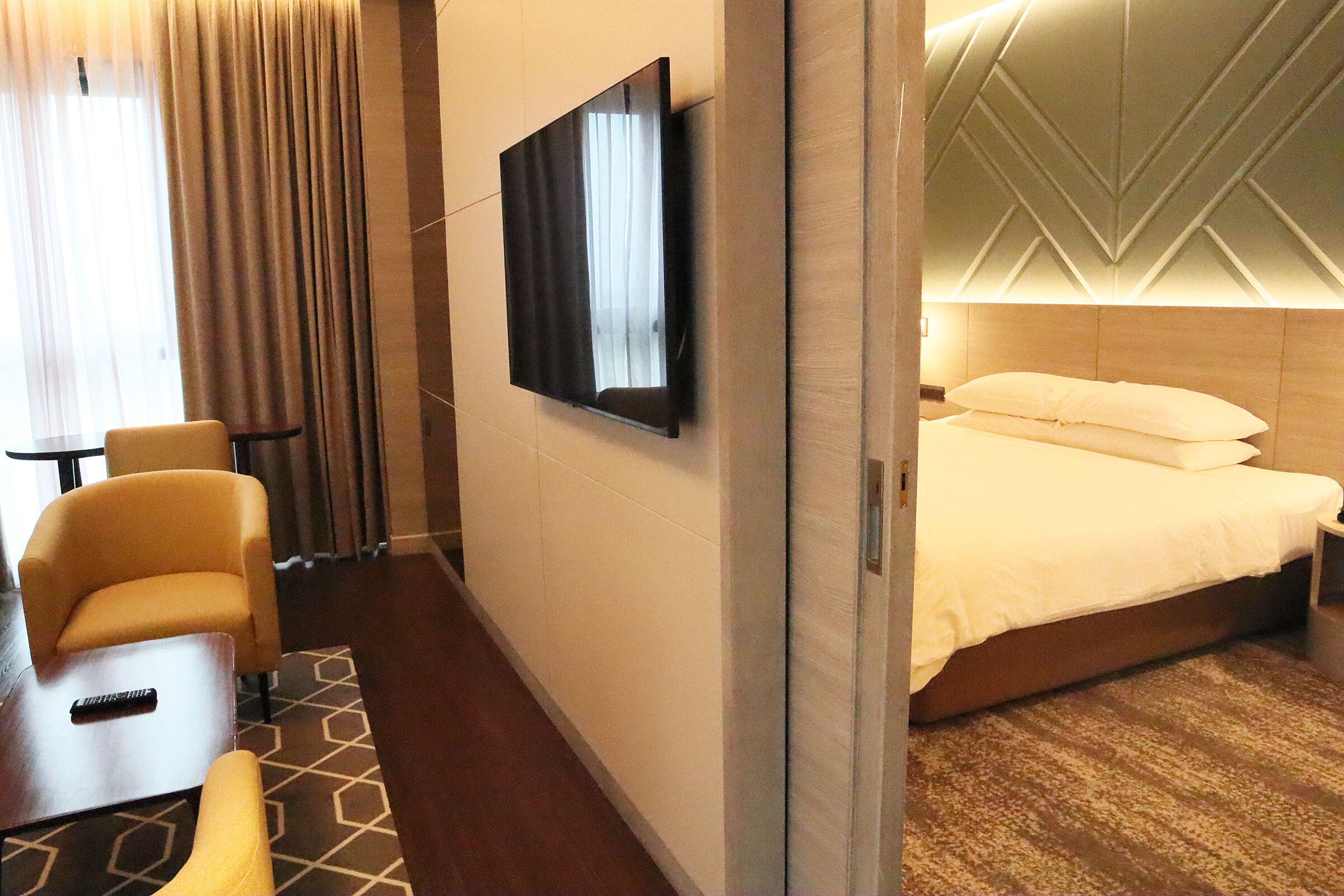 Ein Hotelzimmer mit einem Bett, einem an der Wand montierten Fernseher, einer Sitzecke mit Stuhl und Tisch sowie einem gemusterten Teppich.