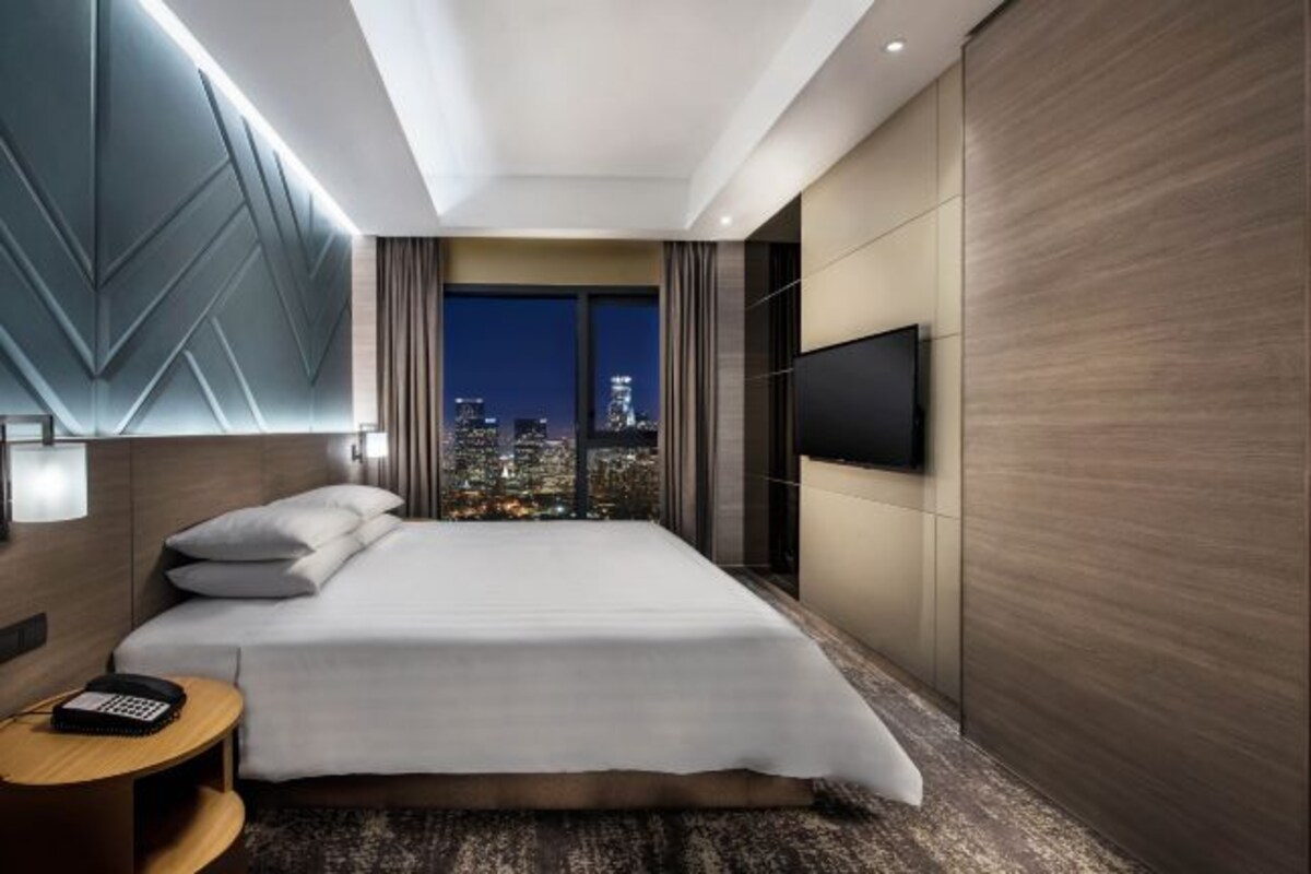 A modern hotel room with a large bed, a city view, a flat-screen TV, and a small side table.