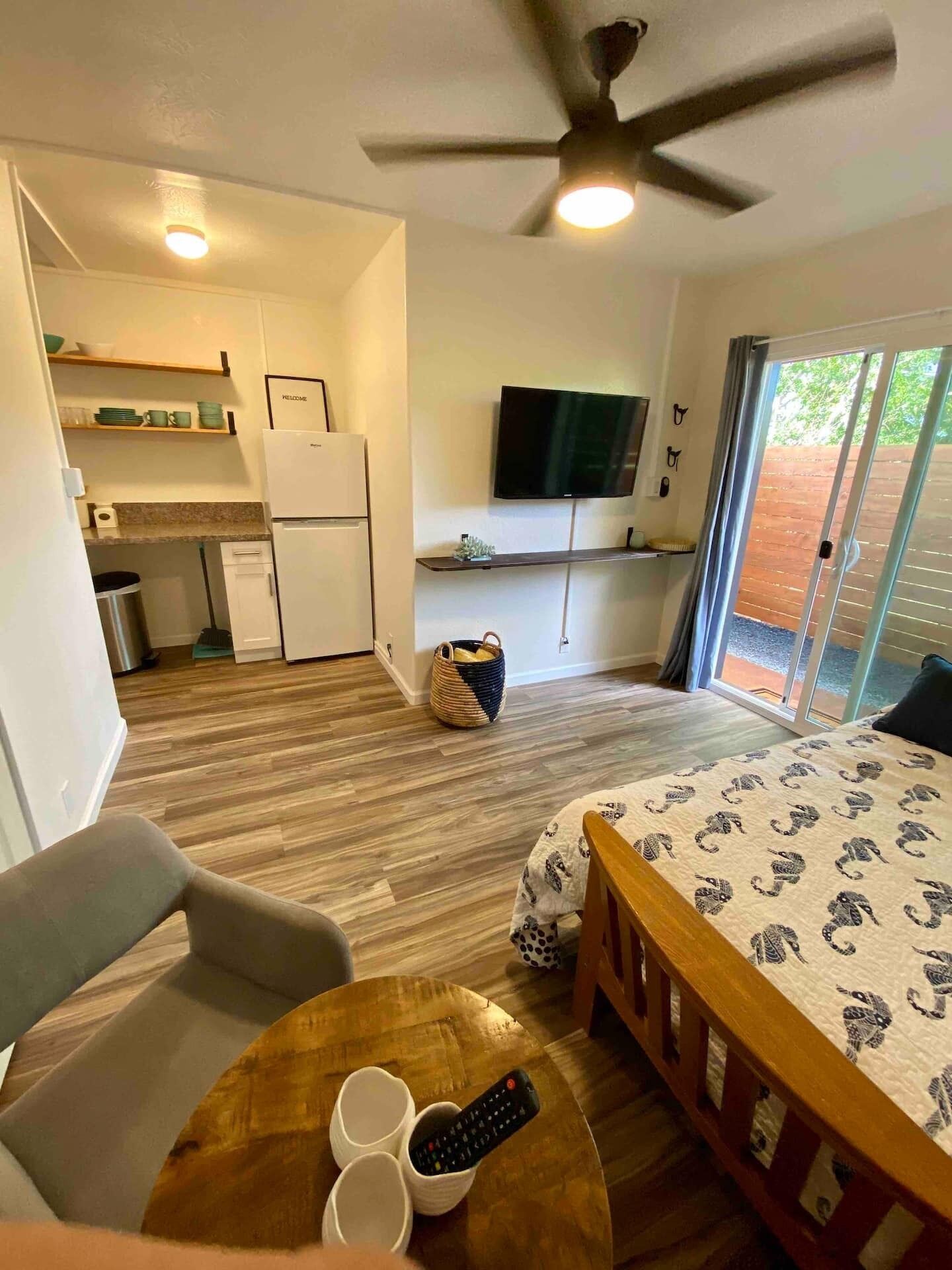 A compact room with a bed, a small table with two cups and a remote, a ceiling fan, a TV on the wall, and a sliding glass door leading to an outdoor area.