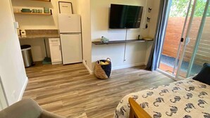 A compact room with a bed, a small table with two cups and a remote, a ceiling fan, a TV on the wall, and a sliding glass door leading to an outdoor area.