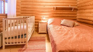Une chambre à coucher dans une cabane en rondins, avec un berceau, une table de chevet avec une lampe, un lit recouvert d’une couverture à motifs et une fenêtre avec des rideaux.
