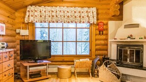 L’intérieur chaleureux d’un chalet en bois comprend une télévision, une cheminée et de grandes fenêtres offrant une vue sur les arbres enneigés.