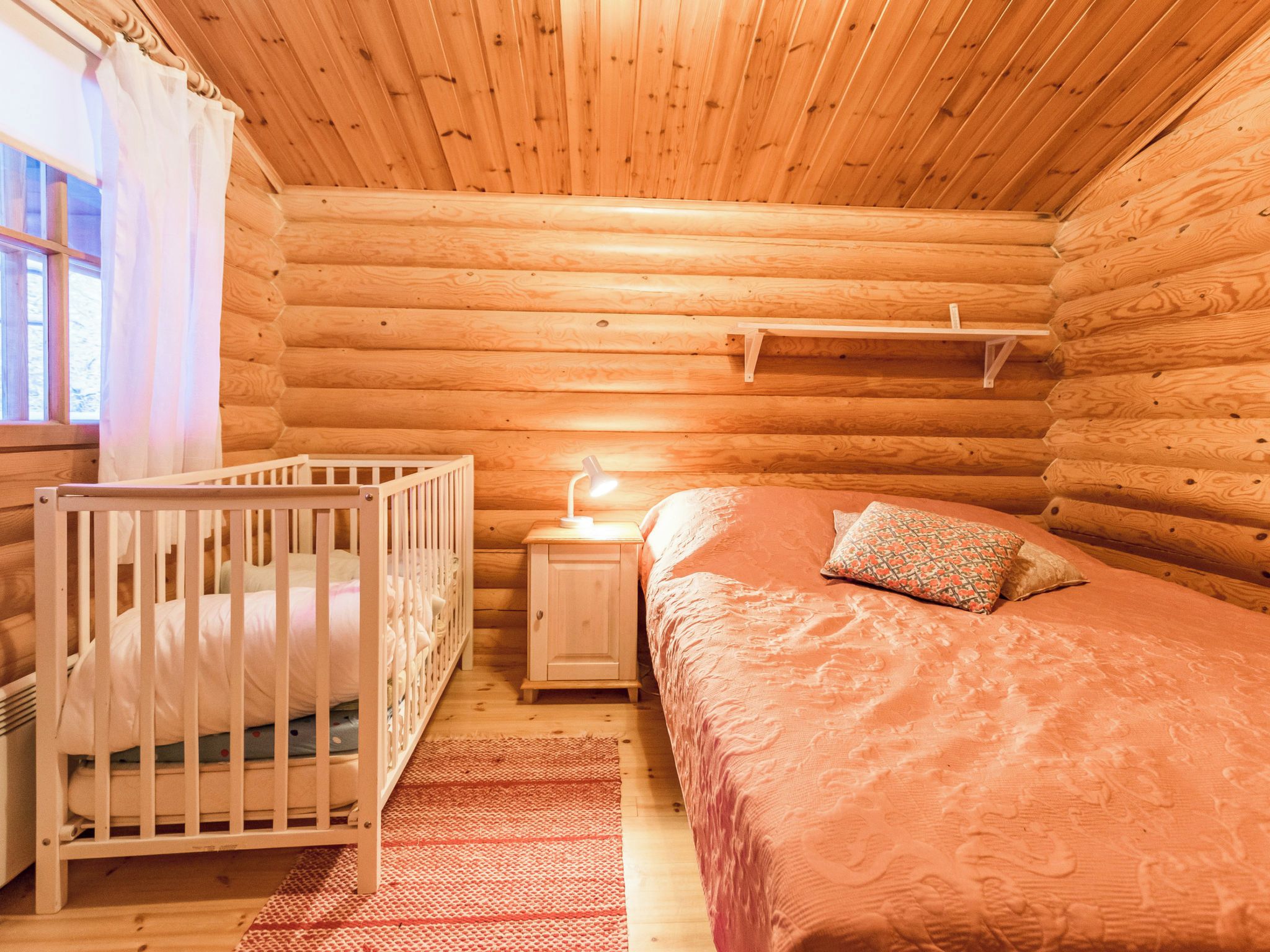 A cozy log cabin bedroom with a crib, a bedside table with a lamp, and a bed with a patterned blanket.
