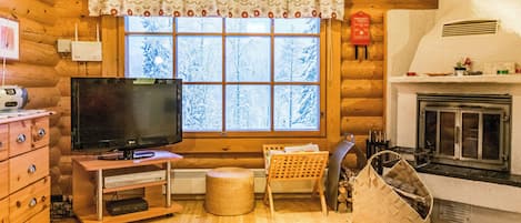 A log cabin interior with a television, wooden furniture, a fireplace, and a window with a view of snow-covered trees.