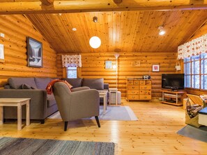 L’intérieur d’une cabane en rondins, avec un canapé, un fauteuil, une commode en bois, un téléviseur et une fenêtre avec des rideaux.
