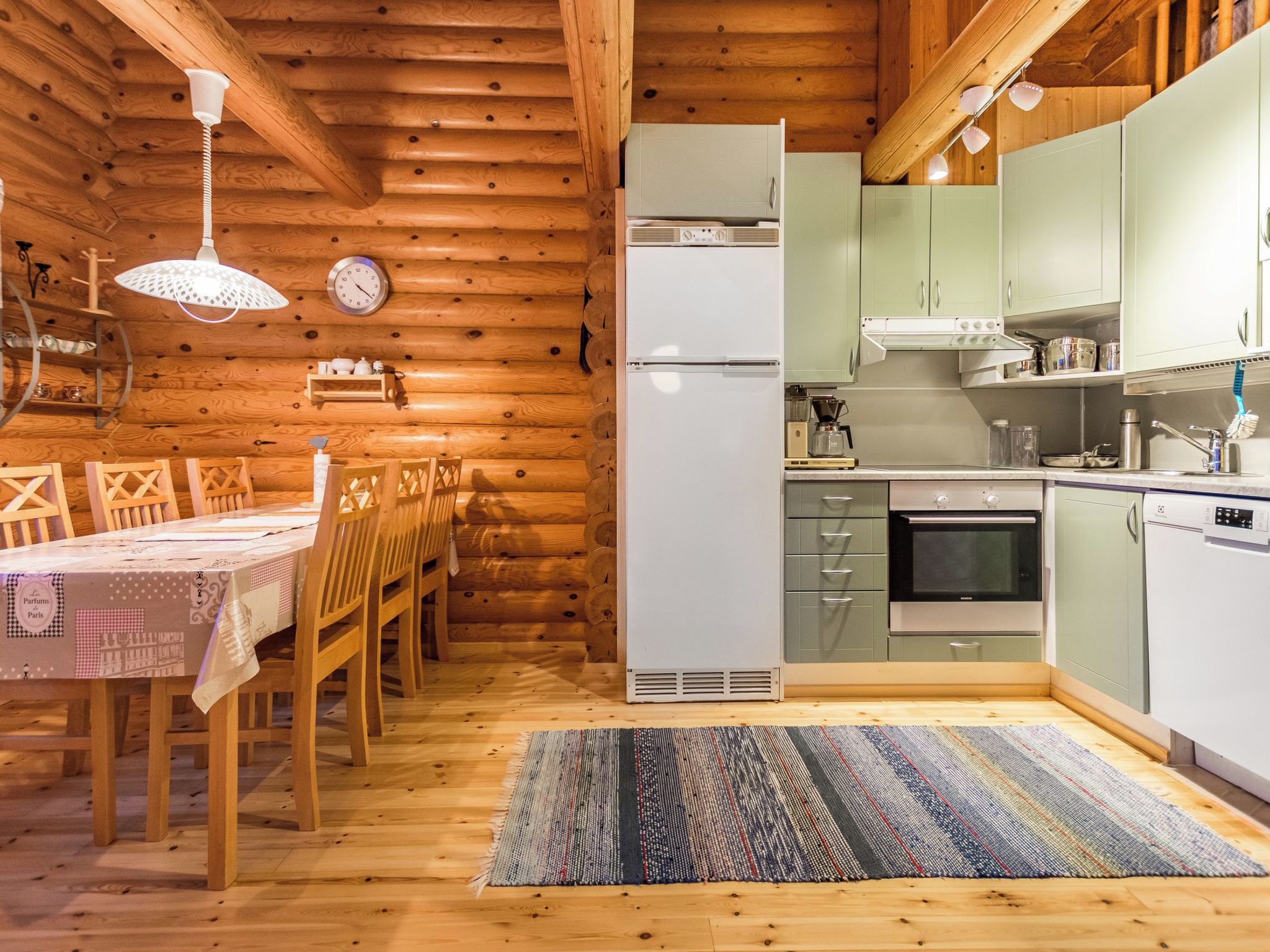 A wooden interior with a dining table, chairs, a kitchen with appliances, and a clock on the wall.