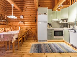 A wooden interior with a dining table, chairs, a kitchen with appliances, and a clock on the wall.