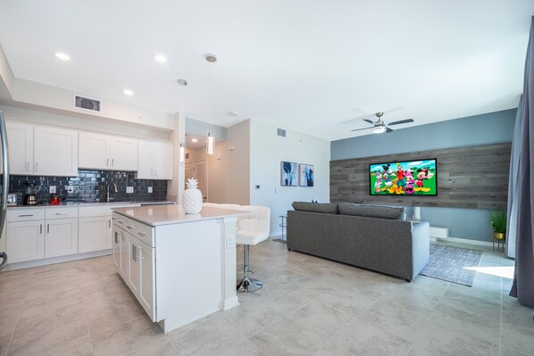 A modern kitchen with white cabinetry, a central island, and a dining area with a grey sofa. - JJ102 Storey Lake Condo 2 bed 2 bath Free Waterpark (Kissimmee)