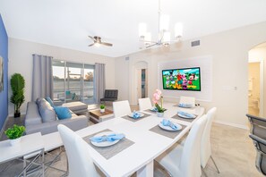 A modern dining area with a large table set for six, white chairs, and a television mounted on the wall displaying cartoon characters.