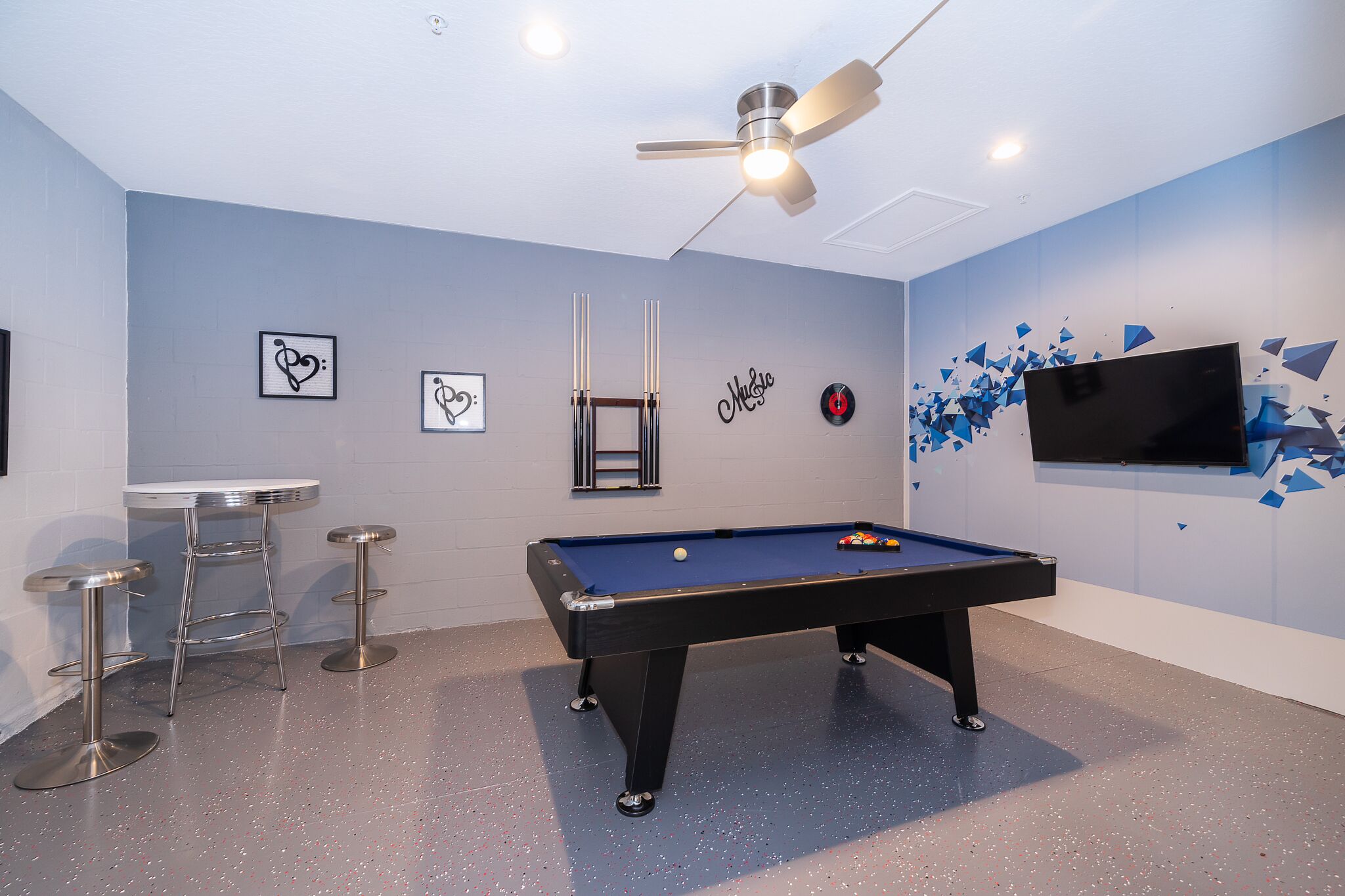 Une table de billard, un ventilateur de plafond, deux tabourets de bar et un téléviseur fixé au mur.