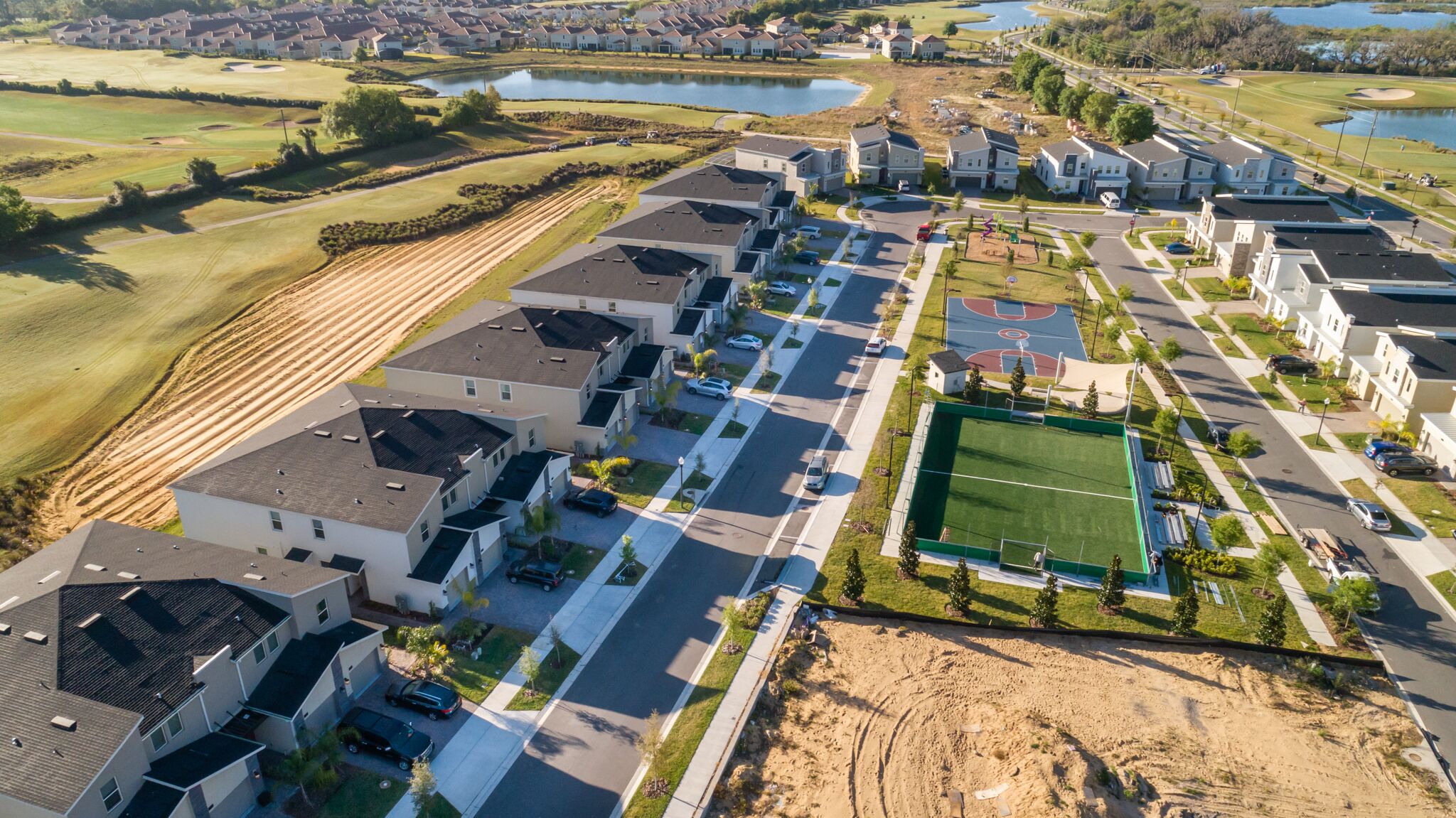 A residential area with modern houses, a sports field, and a lake in the background.
