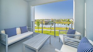 A balcony with a white sofa and chairs, a table, and a view of a water body and buildings.