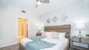 Une chambre d’hôtel avec un grand lit, une table de chevet avec une lampe, un ventilateur de plafond et des œuvres d’art décoratives au mur.