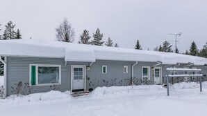 A single-story, grey wooden cabin with a snow-covered roof, a front door, and a small porch area.