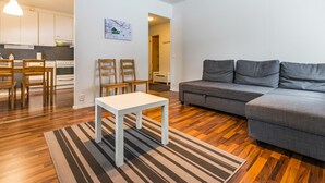 A modern living room with a grey sofa, a white table, and striped rugs. There is a kitchen area with wooden cabinets and a dining table with chairs.