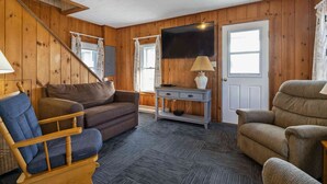 Una sala de estar acogedora con paredes de madera, una escalera, un televisor de pantalla plana, un sofá, un sillón mecededor y una lámpara.