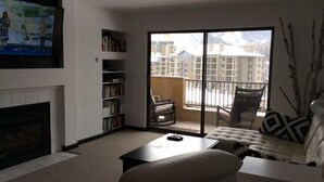 A living room with a fireplace, a sofa, a coffee table, and a bookshelf. There is a large window with a view of buildings and a balcony. - Steps from Gondola, views of Beaver Creek slopes, newly remodeled 2 BR 2BA Condo (Avon)