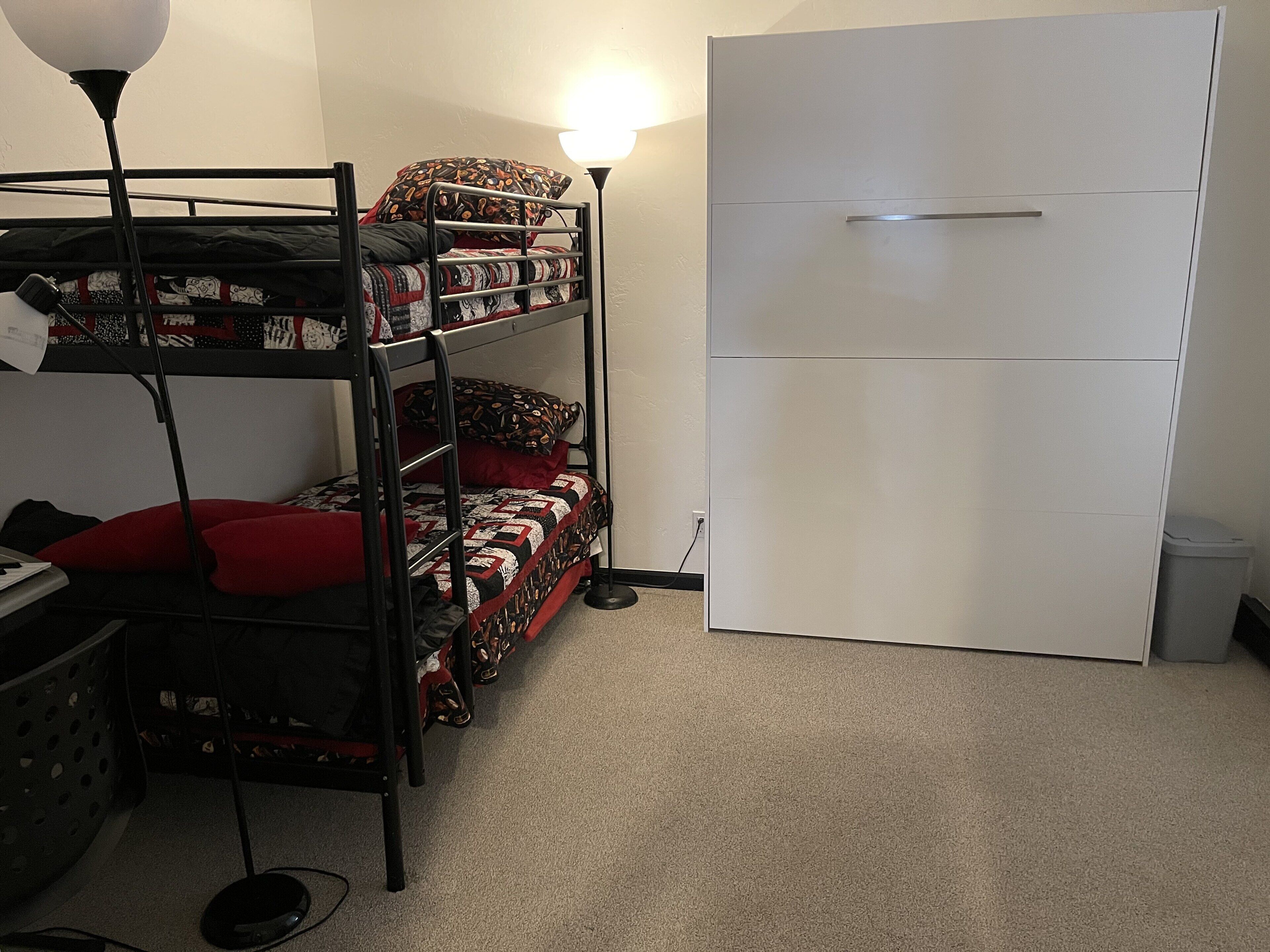 A bunk bed with patterned bedding, a white wardrobe, and a desk with a lamp.