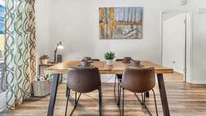 A dining area with a wooden table, two brown chairs, a potted plant, and a painting on the wall. - Stylish Pool Home w/ Grill by GNV's Best Dining! (Gainesville)
