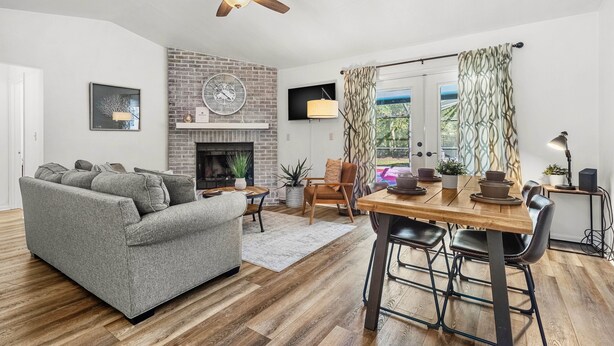 A modern living room with a grey sofa, a wooden dining table with chairs, a brick fireplace, and a ceiling fan. - Stylish Pool Home w/ Grill by GNV's Best Dining! (Gainesville)