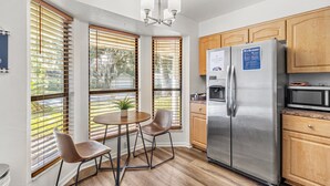 A modern kitchen with a stainless steel refrigerator, wooden cabinets, and a dining area with two chairs and a table. - Stylish Pool Home w/ Grill by GNV's Best Dining! (Gainesville)