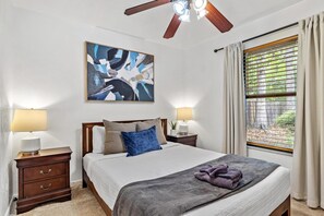 Une chambre à coucher avec un lit, deux tables de chevet, un ventilateur de plafond, une fenêtre avec des rideaux et un tableau au mur.