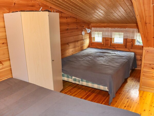 Un pequeño dormitorio de cabaña de madera con una cama individual, un armario y una ventana con cortinas de encaje.