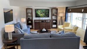 Una sala de estar moderna con un sofá gris, una mesita de centro y un televisor. Hay dos mesitas de noche con lámparas, una estantería y una ventana con persianas.