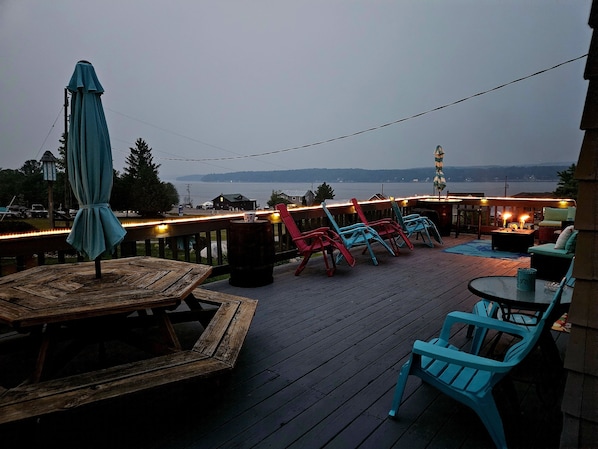 A deck with wooden furniture, a view of the water, and string lights.