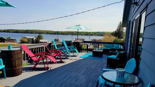 A wooden deck with colorful chairs and a table, overlooking a lake with houses in the background. - Shore Beats Work (East Jordan)