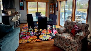 A dining area with a table and chairs, a colorful rug, and a view of the outdoors. - Shore Beats Work (East Jordan)
