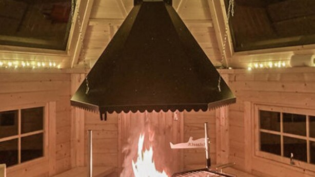 Interior de una cabaña de madera con chimenea central, paredes de madera y techo inclinado.