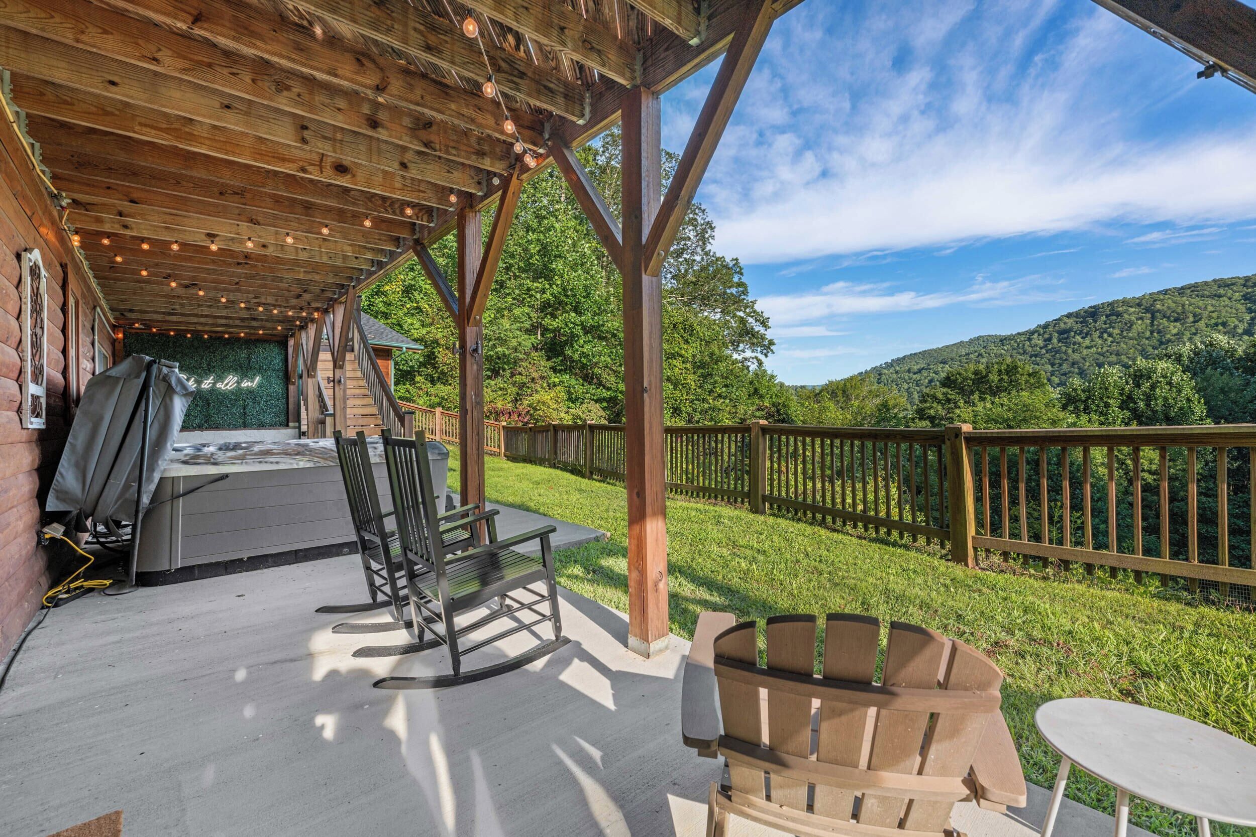 An outdoor patio with wooden furniture, a hot tub, and a view of a forested hillside.