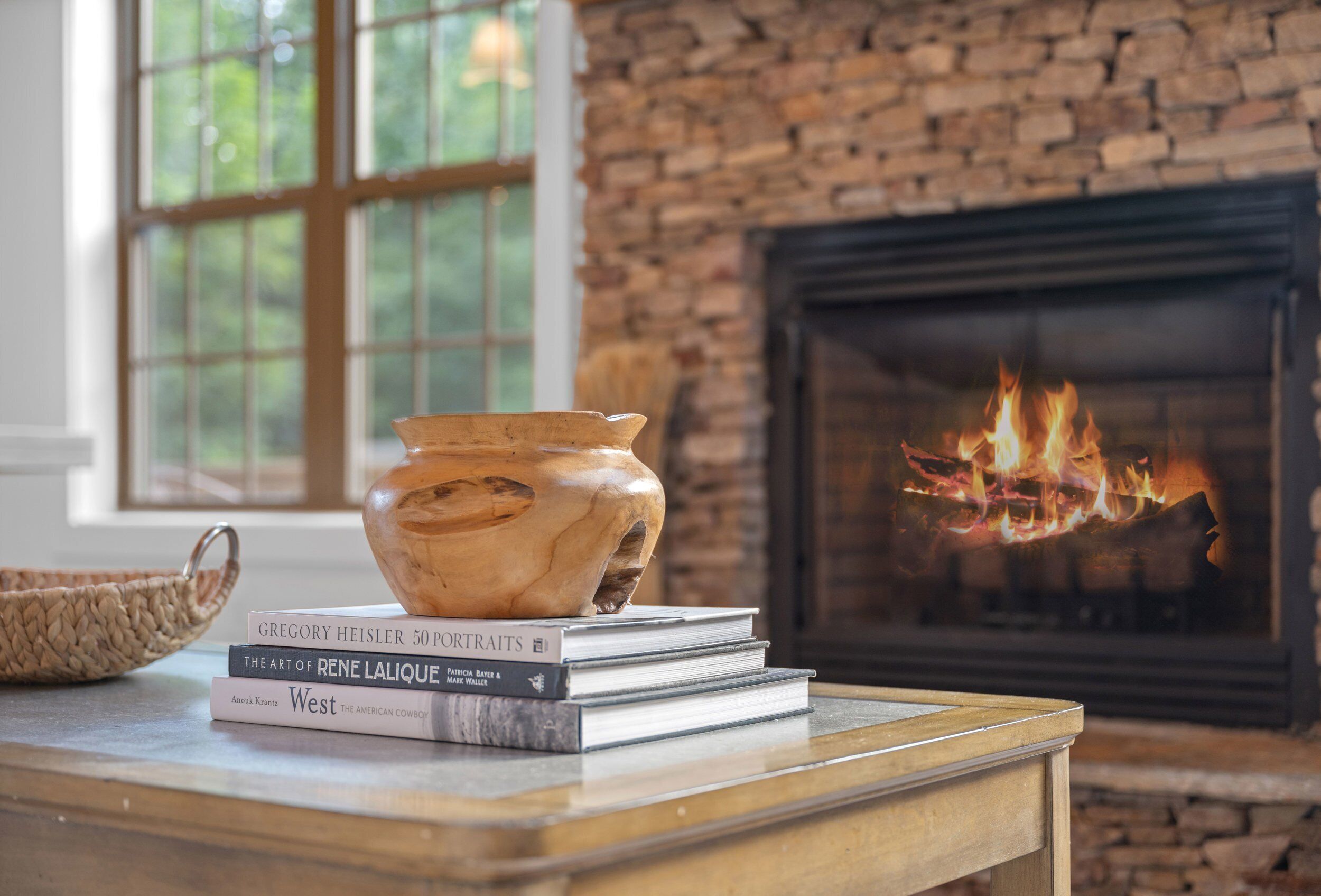 A fireplace with a fire, a wooden table with a vase and books, and a brick wall with a window.