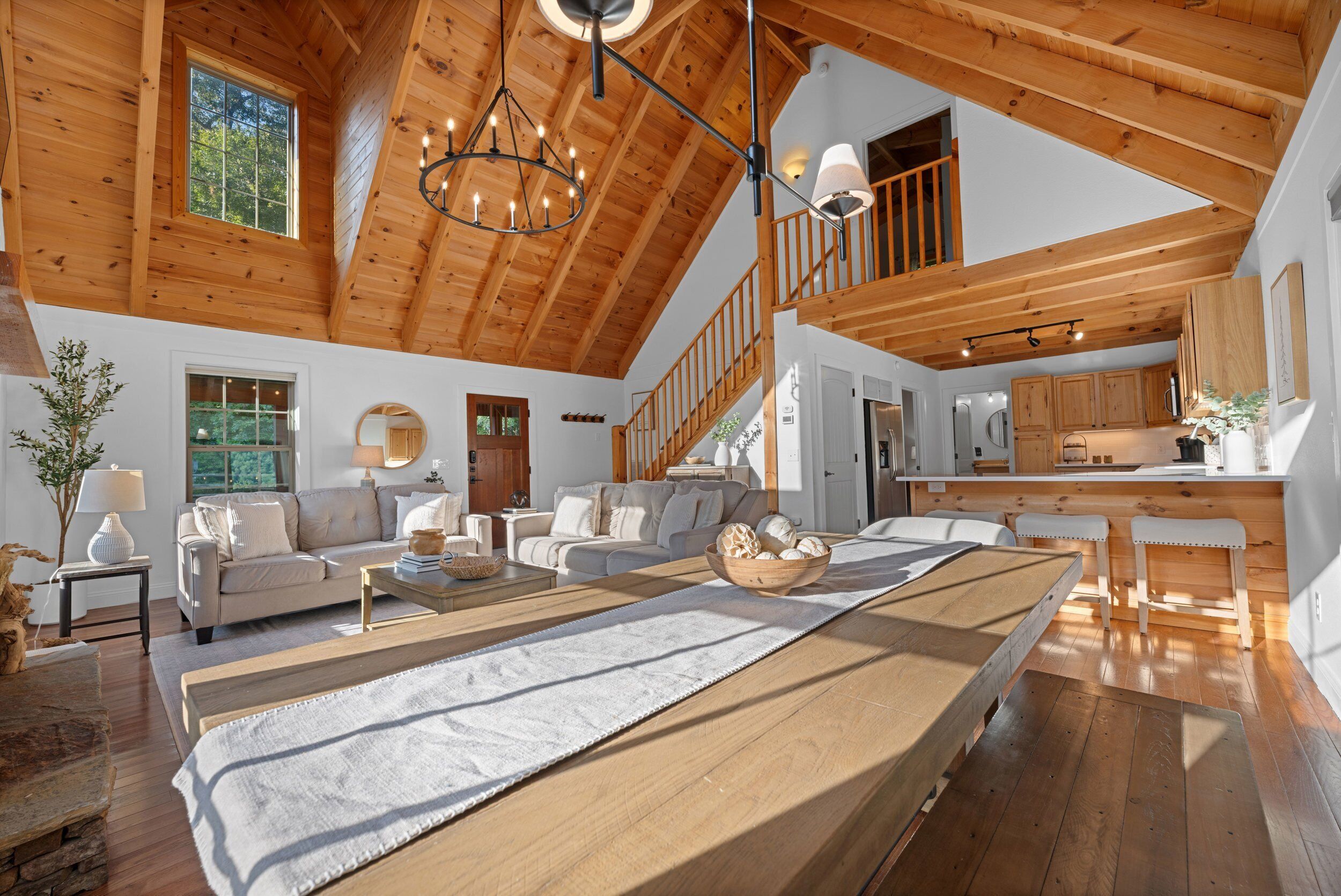 A spacious living area with a wooden ceiling, a sofa set, a dining table, and a kitchen in the background.