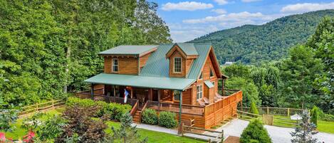 A log cabin with a porch, surrounded by a fenced yard and a mountain backdrop.