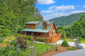 A log cabin with a porch, surrounded by a fenced yard and a mountain backdrop. - Vista View - mountain views, hot tub, game room, fire pit, fenced in yard (Sugar Grove)