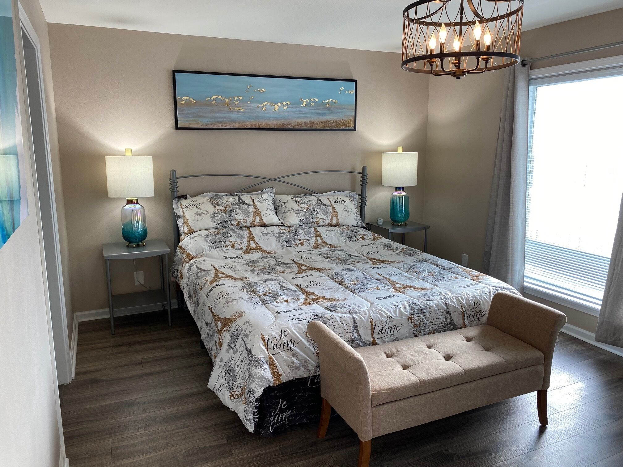 A bedroom with a bed, two bedside lamps, a bench, a painting, and a window with blinds.