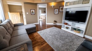 A living room with a grey sectional sofa, a flat-screen TV, a grey rug, wooden flooring, a white entertainment unit, and a door leading to another room. - The Elowen Suite (Drumheller)