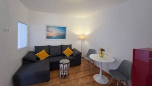 Ein modernes Wohnzimmer mit einem grauen Sofa, zwei gelben Kissen, einem kleinen Beistelltisch, einem weißen runden Tisch mit zwei Stühlen, einer Stehlampe und einem Gemälde an der Wand.