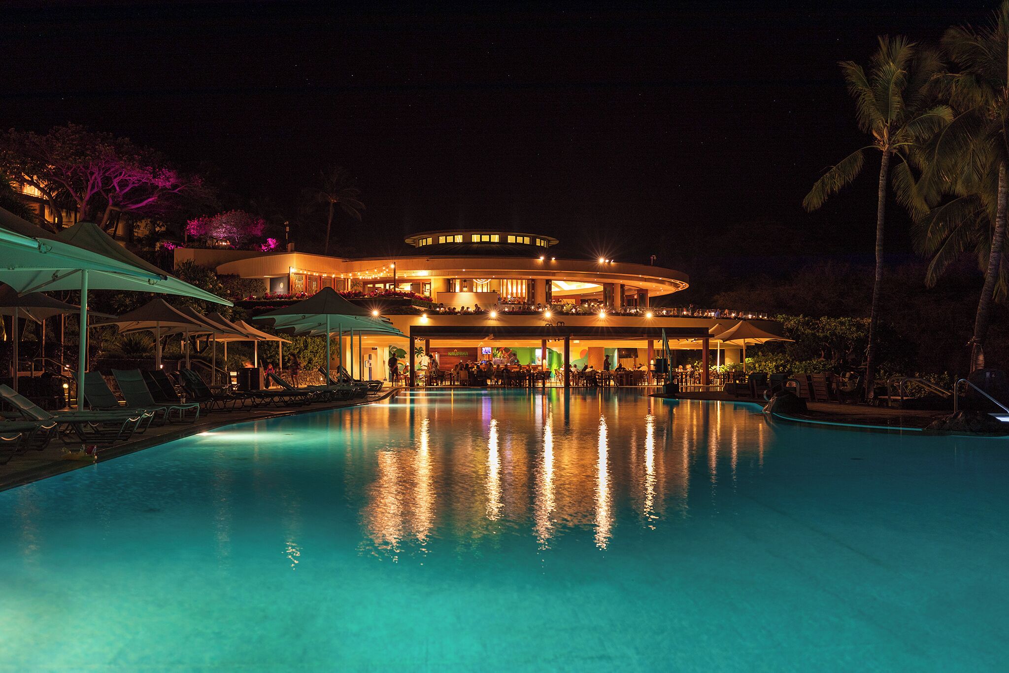 A large, illuminated hotel with a pool in the foreground.
