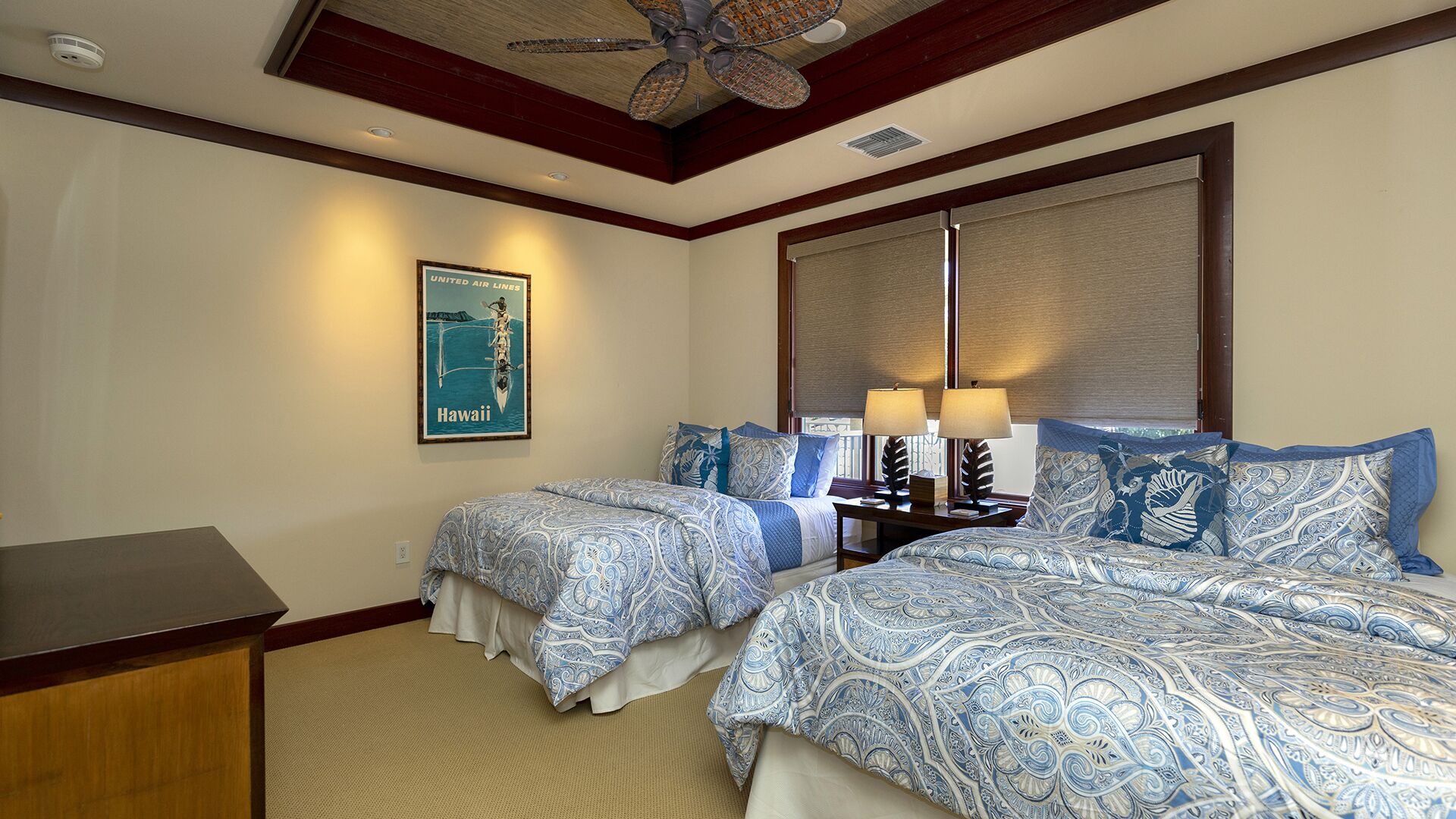 Ein Hotelzimmer mit zwei Betten, einem Schreibtisch, einem Deckenventilator, einem großen Fenster mit Jalousien und einem Hawaii-Poster.