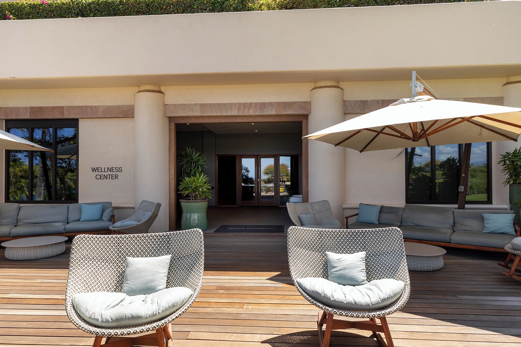 Un espace extérieur aménagé avec des chaises en osier et un grand parasol, une enseigne indiquant un centre de bien-être, et une terrasse en bois.