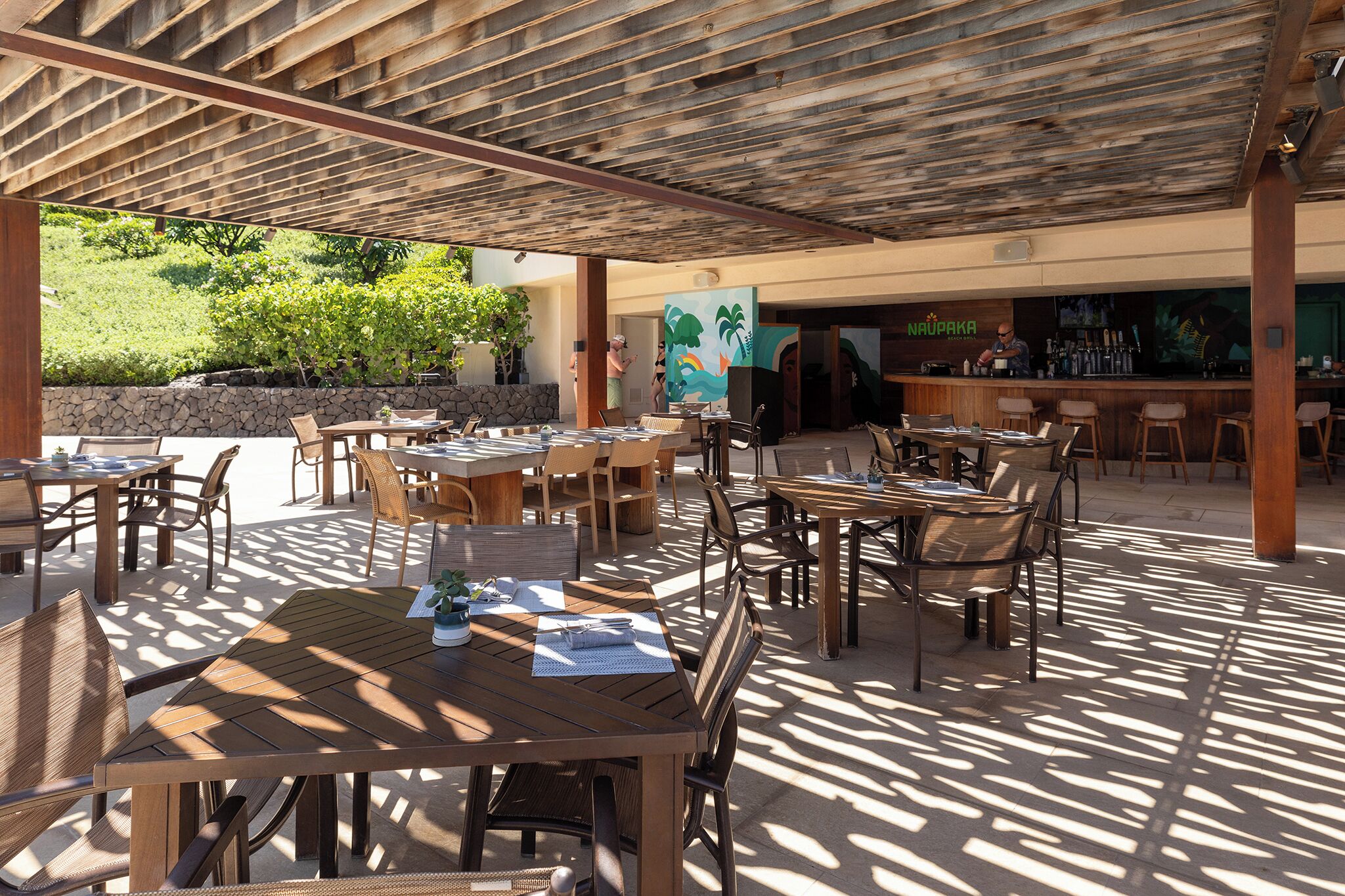 An outdoor patio area with wooden tables and chairs, a stone wall, and a bar area with a mural.
