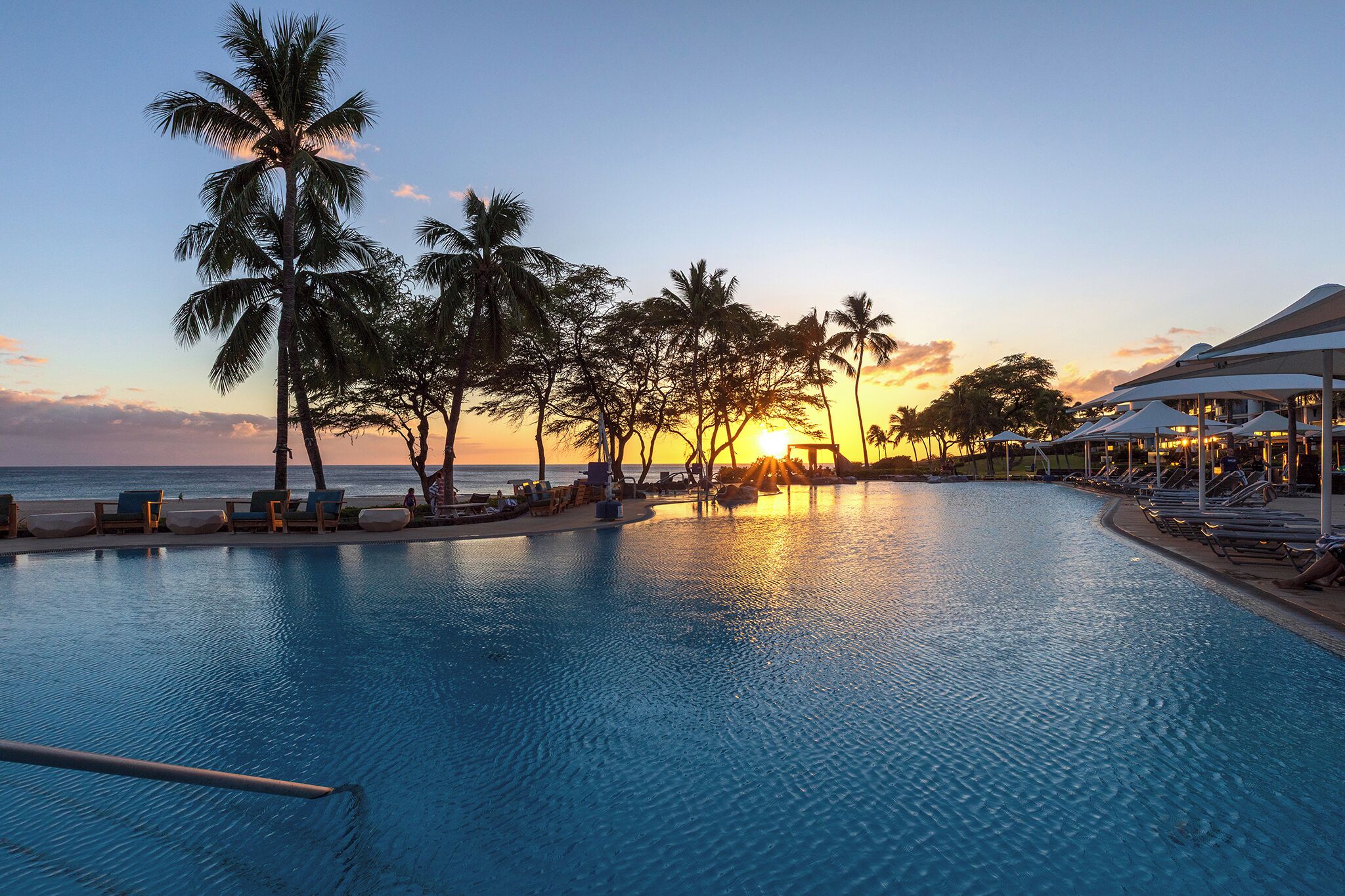A poolside area with lounge chairs and umbrellas, palm trees, and a sunset view.