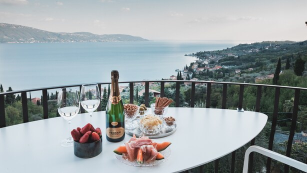 Ein Tisch mit einer Weinflasche, Gläsern, Erdbeeren und einer Speisenplatte auf einem Balkon mit schöner Aussicht.