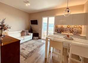 A modern living room with a sofa, coffee table, and a TV. There's a kitchen area with a tiled backsplash and a dining table.