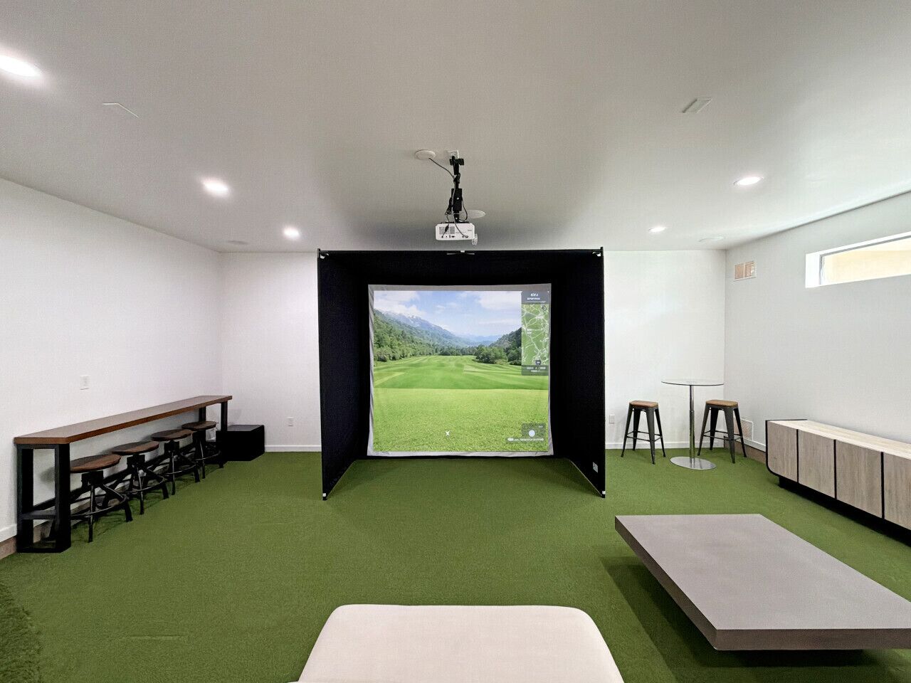 Ein Zimmer mit großem Bildschirm, der einen Golfplatz zeigt, mit grünem Teppich, einem langen Tisch und einem an der Decke befestigten Projektor.