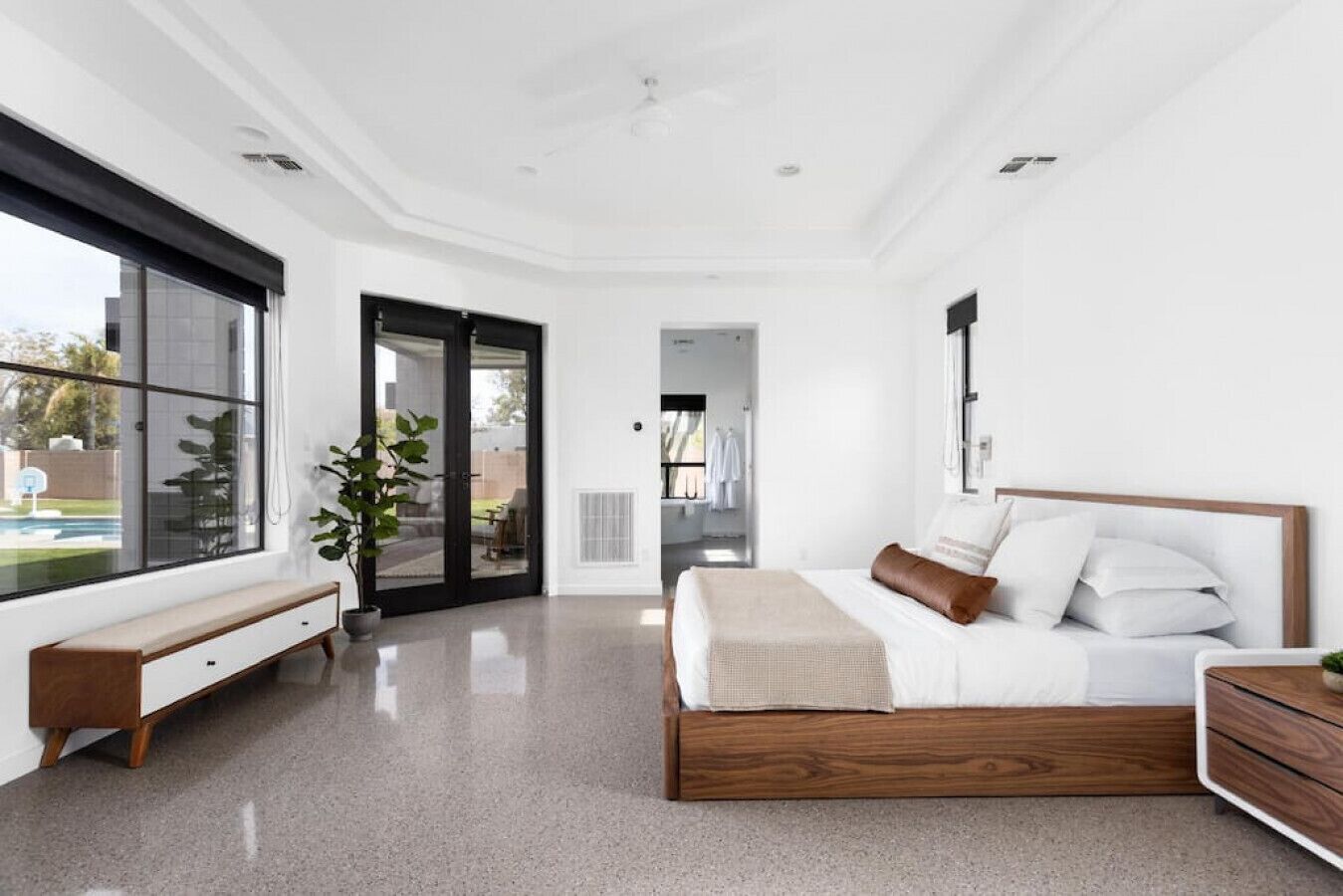 A modern bedroom with a large bed, a bench, and a view of a backyard through a sliding door.